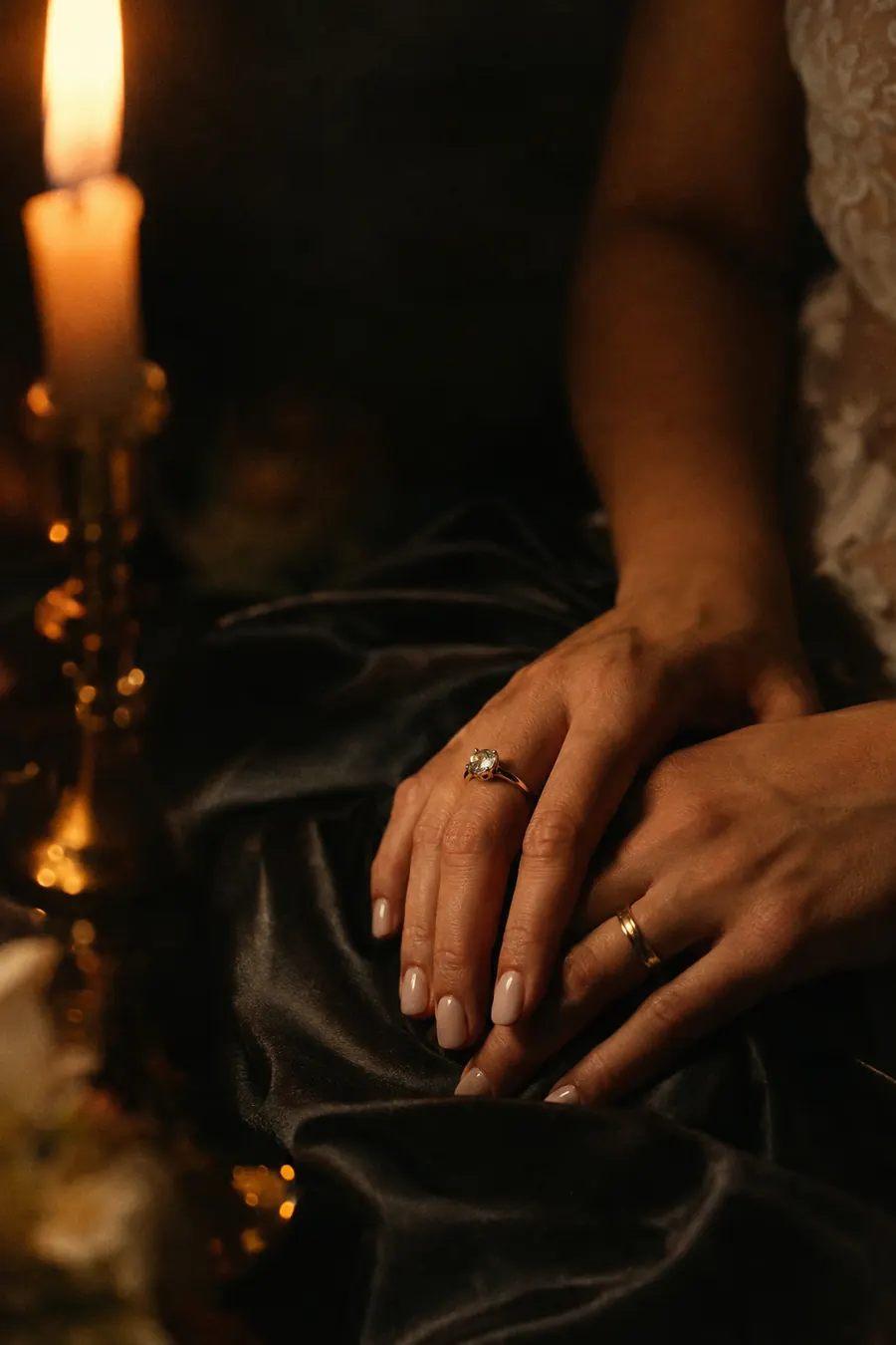 A bride's hands with gold rings resting on dark silk, lit by candlelight